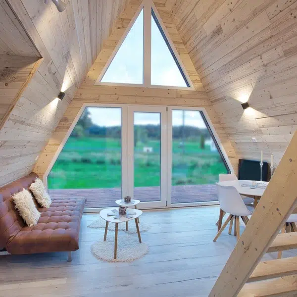 Salon du chalet A-Frame Iorik avec baie vitrée triangulaire pleine hauteur, canapé en cuir et vue sur la nature vosgienne, près de Gérardmer
