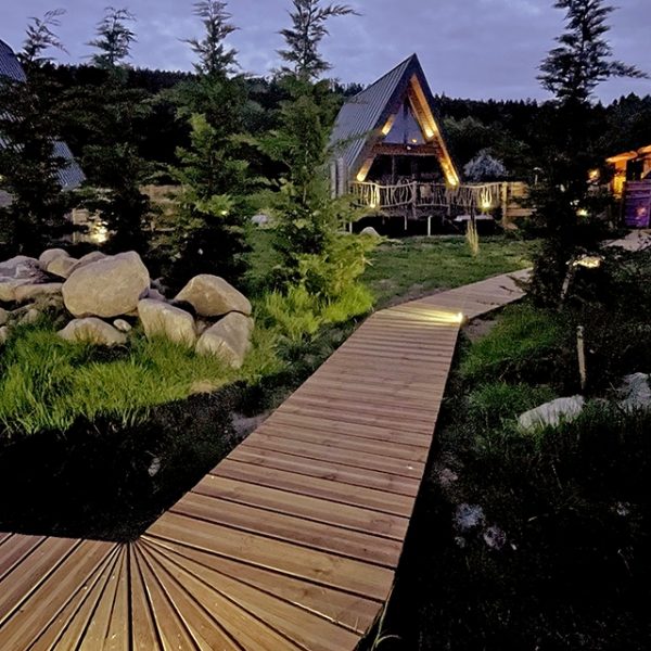 Chalet A-Frame Iorik illuminé de nuit, allée en bois menant à la terrasse, niché en forêt à 10 min de Gérardmer dans les Vosges